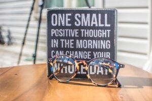 Piccolo riquadro su un tavolo e dietro a degli occhiali, con la scritta che recita "One small positive thought in the morning can change your whole day"